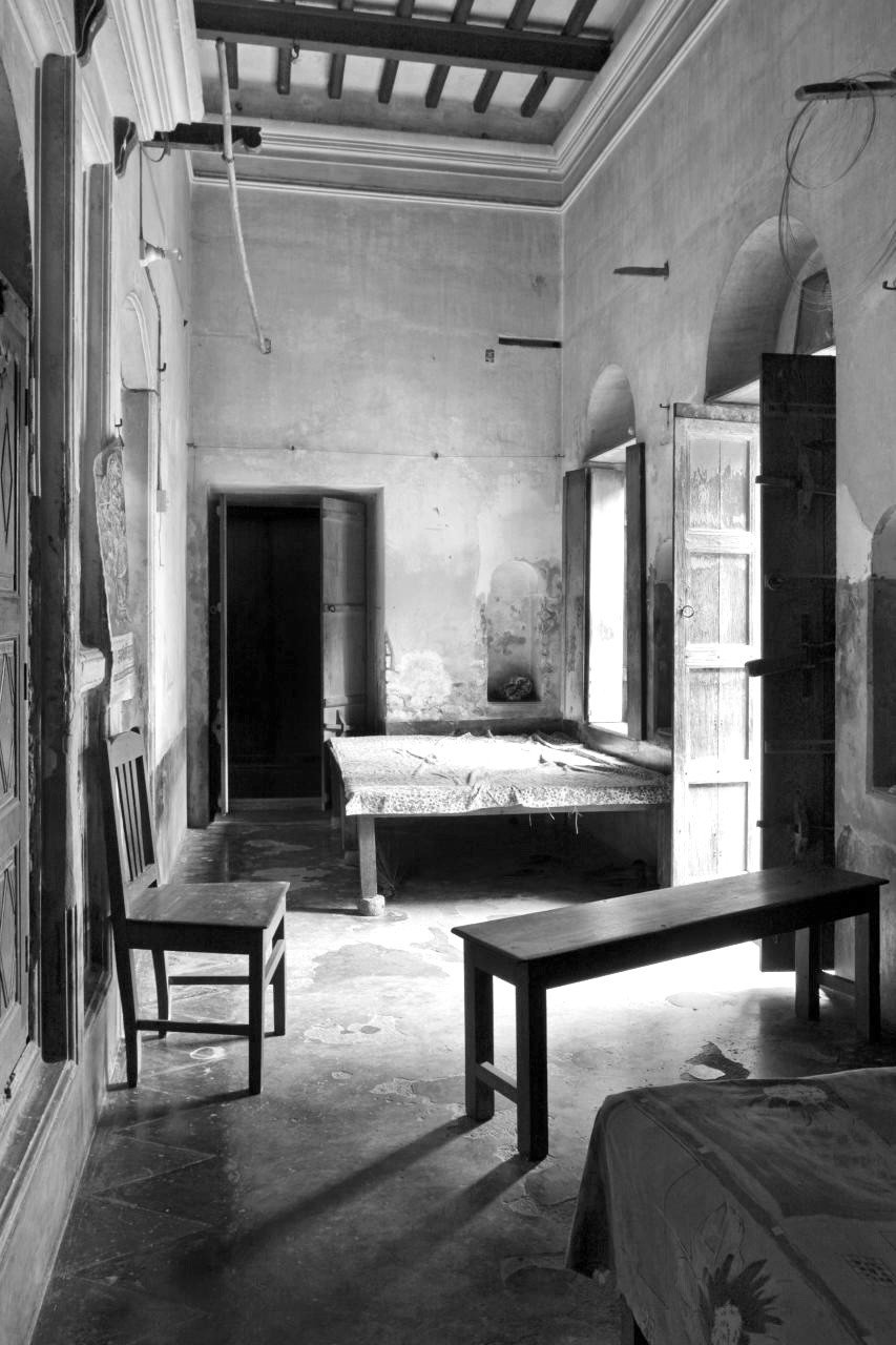 Black and white shot of simple room with arched door way, tile floor, wooden chair and bench, and flat wooden bed frame