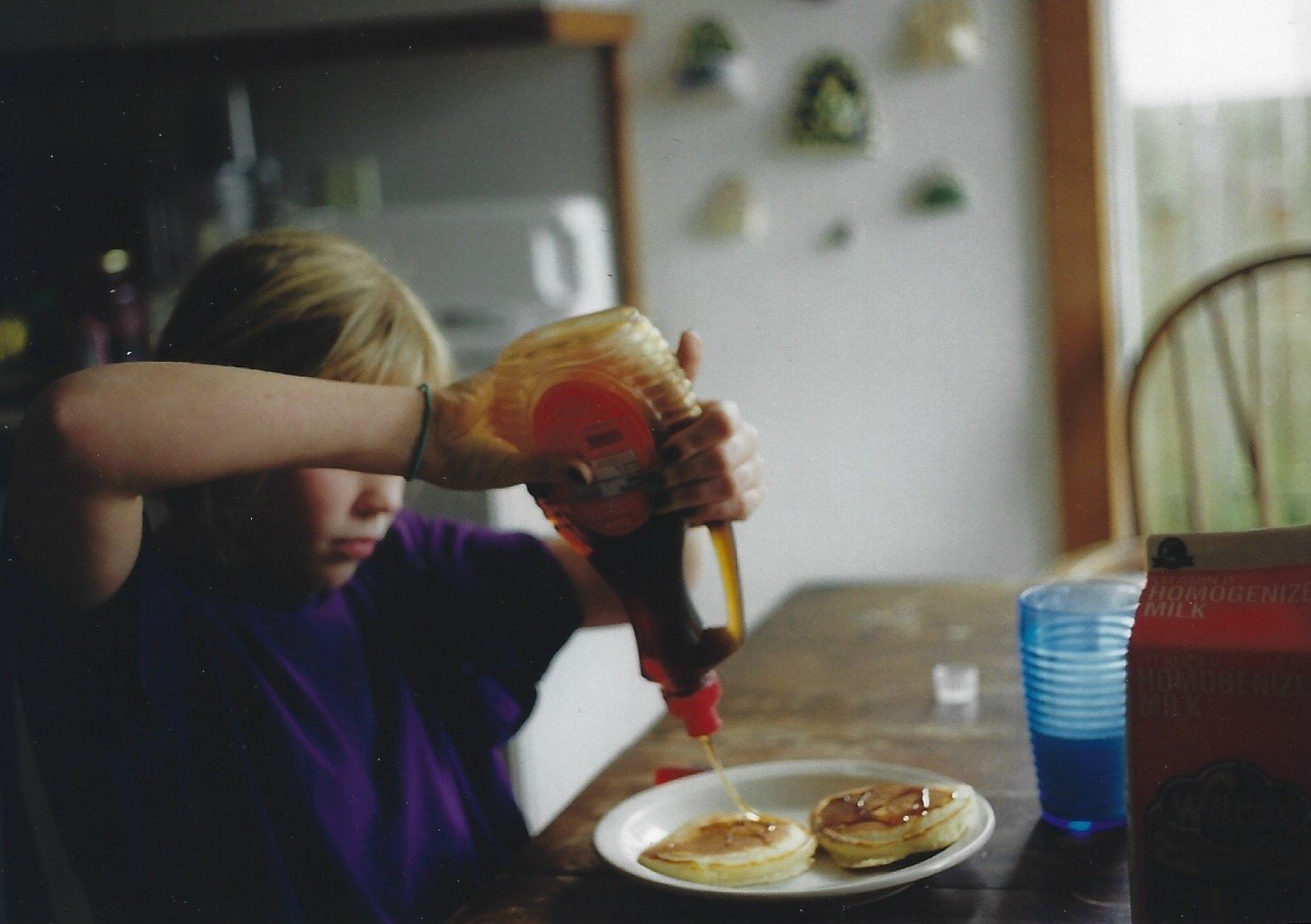 Childhood photo of blonde girl pouring syrup onto pancakes at kitchen table