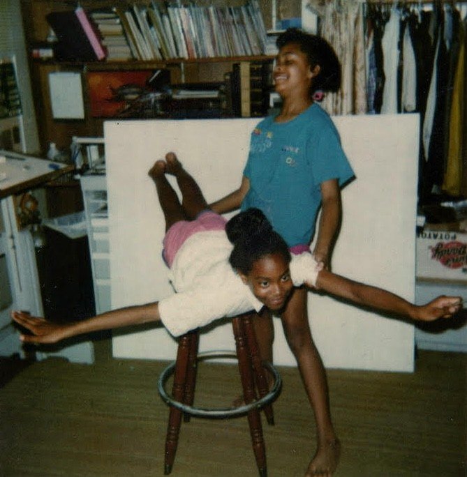 Kenturah Davis and her sister as teenagers, one balancing with her stomach on a stool like an airplane, the other standing and holding her up