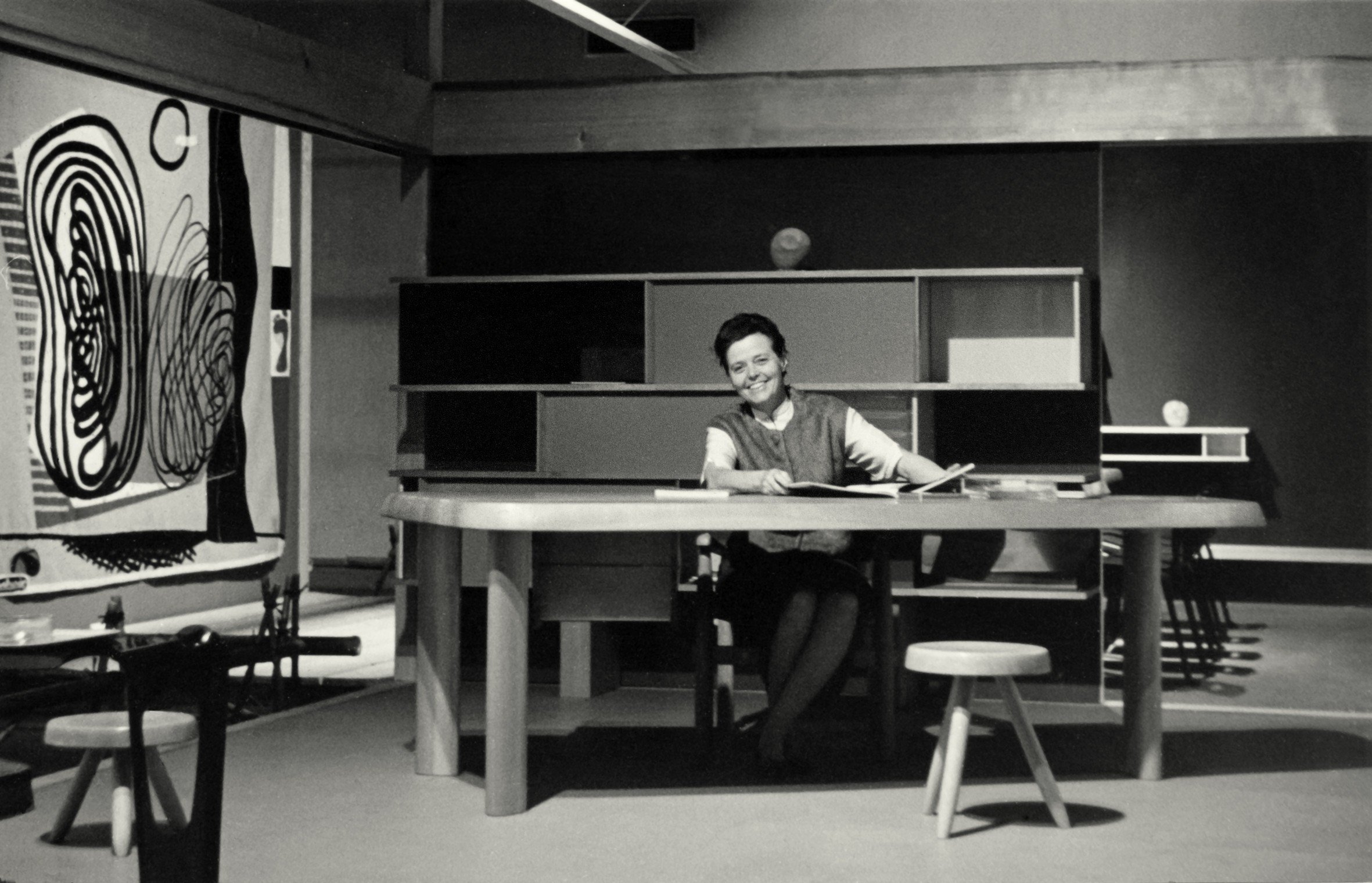 Black and white photo of Charlotte Perriand sitting at desk smiling in the exhibition “Proposition d’une synthèse des arts,” in Tokyo