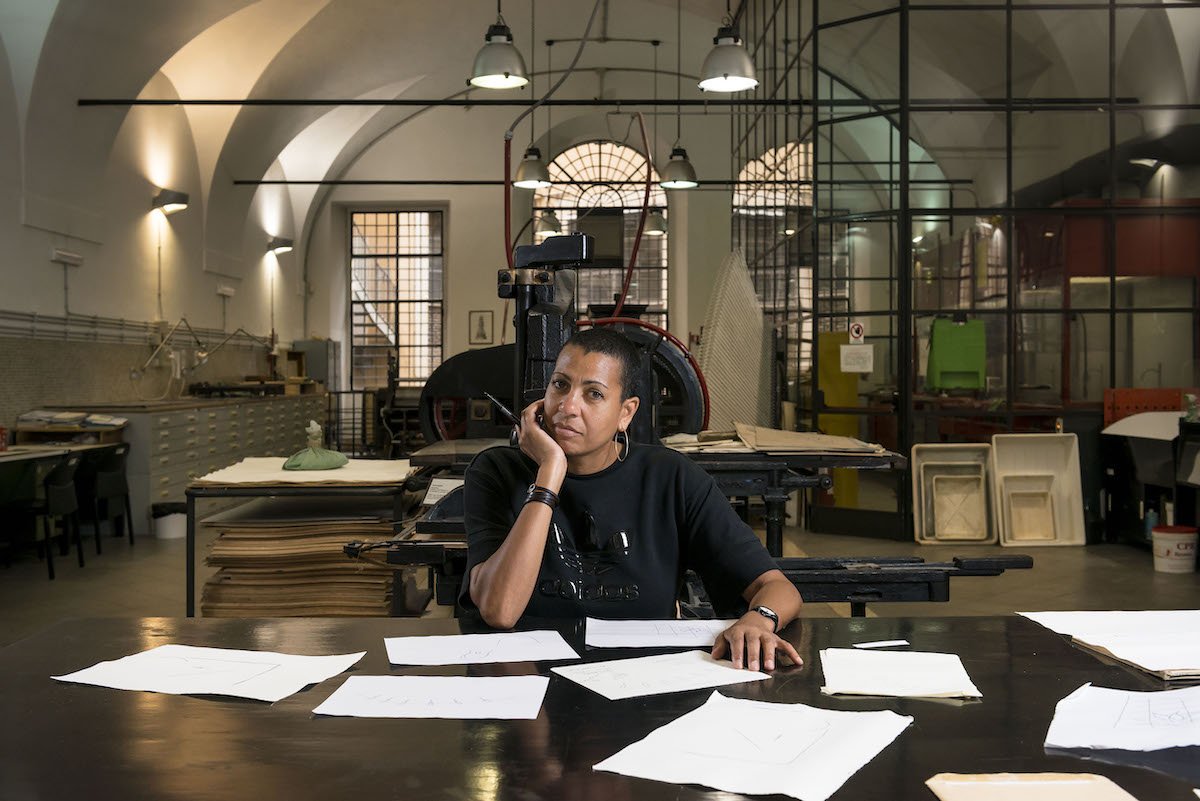 A woman sits at a desk covered with papers in an industial space.