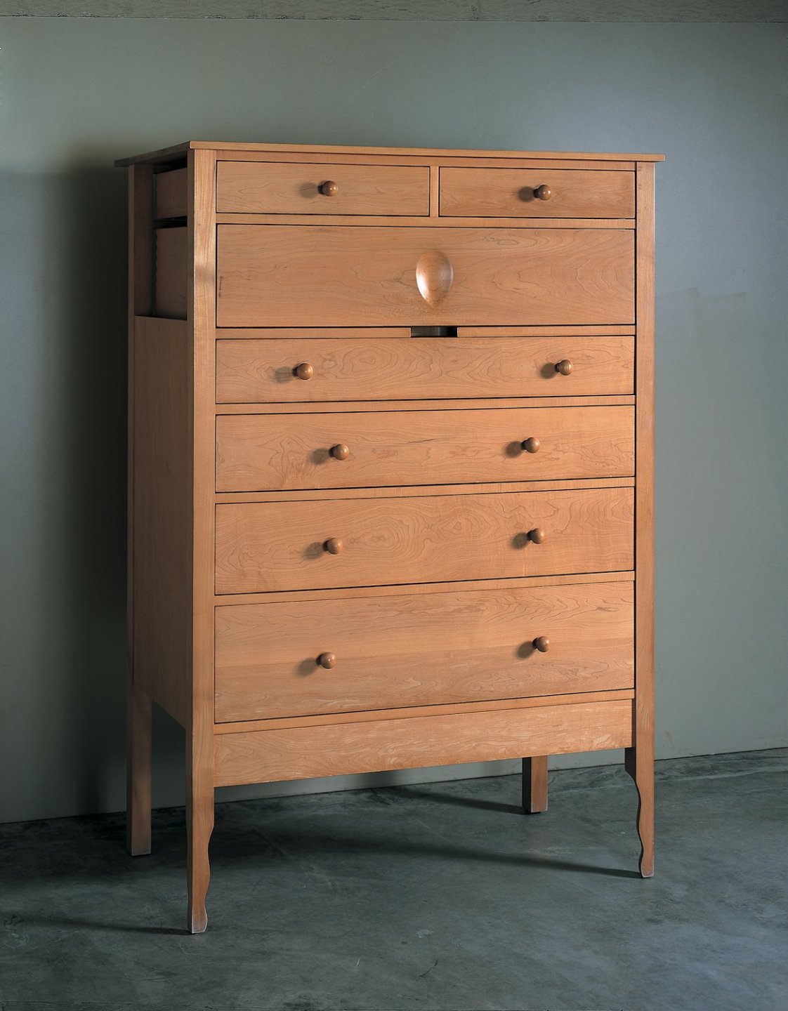 Light brown maple chest of drawers with tear-shaped divot in the middle of the second-to-top drawer against a green-gray background