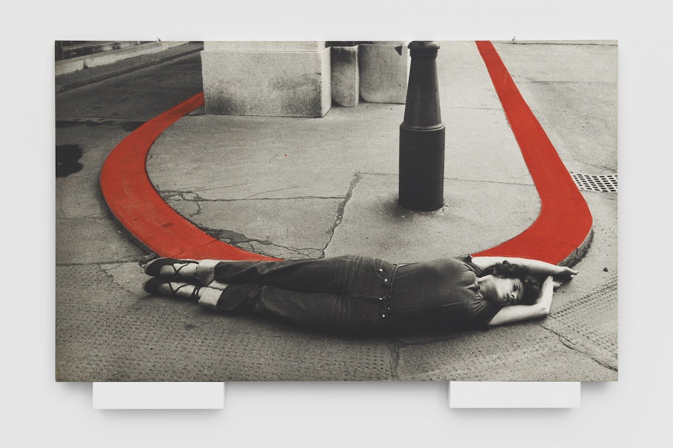 Black and white image of VALIE EXPORT lying on her side against a rounded street gutter, which is colored bright red